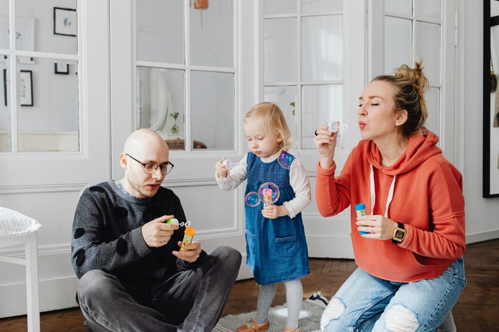 בתמונה רואים ילדה מפריחה בועות סבון עם ההורים שלה. זו דוגמא לפעילות חברתית חושית שמאפיינת את גישת הדנבר. הילדה וההורים במשחק משותף שיכול לעודד לקיחת תורות, חיקוי וקשב משותף. 